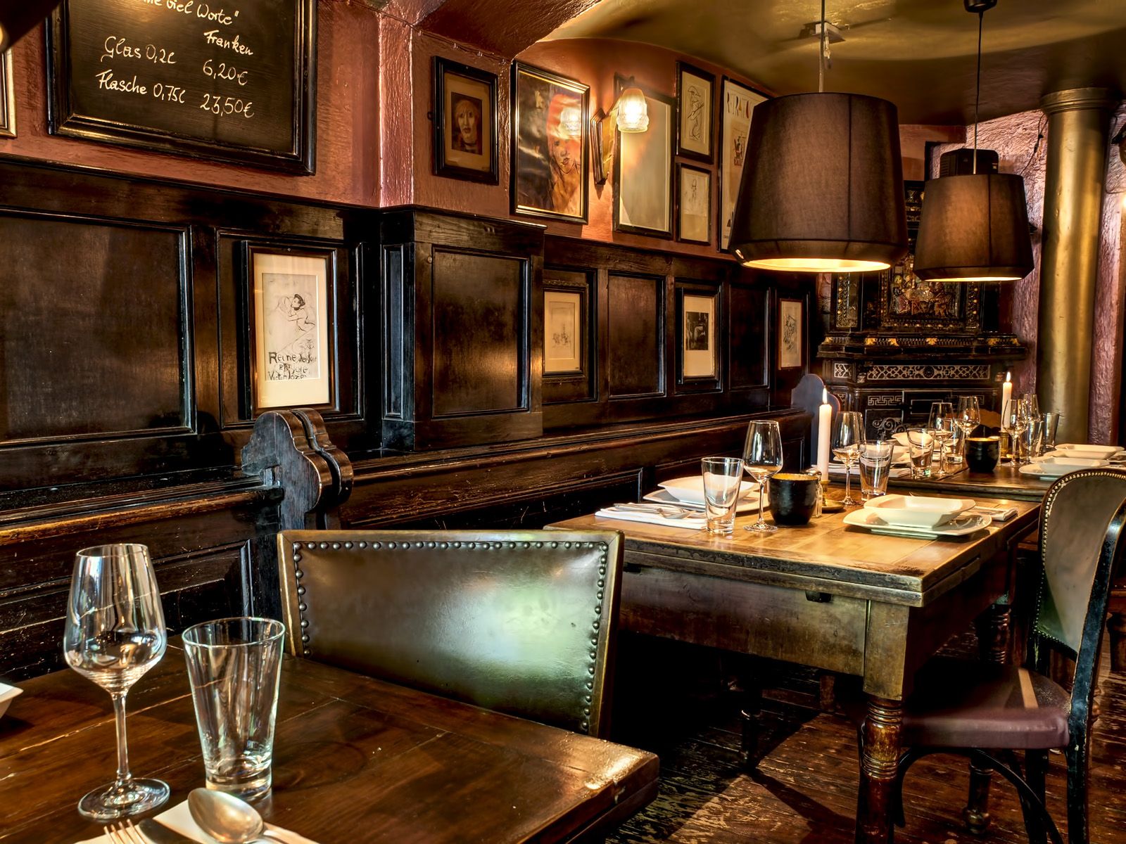 Auf diesem Bild sehen wir einen gedeckten Tisch mit Servietten, Gläsern, weißem Porzellan, Kerzen und silberbesteck. Im Hintergrund befindet sich ein alter Kachelofen im Biedermeier Stil. Dazu sieht man die alten Kirchenbänke sowie zahlreiche Bilder an den Wänden.