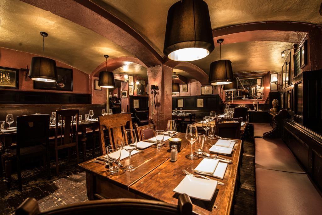 Auf diesem Bild sieht man den vorderen Raum des Restaurants. Im Vordergrund haben wir einen Tisch für vier Personen, dieser ist bereits eingedeckt mit servierten, Besteck, Brottellern, Weingläsern sowie Wassergläsern. Man sieht die alte Holzbank, viele Bilder an den Wänden, große Lampenschirme über den Tischen, die goldene Decke, das Gewölbe, die massiven Säulen.