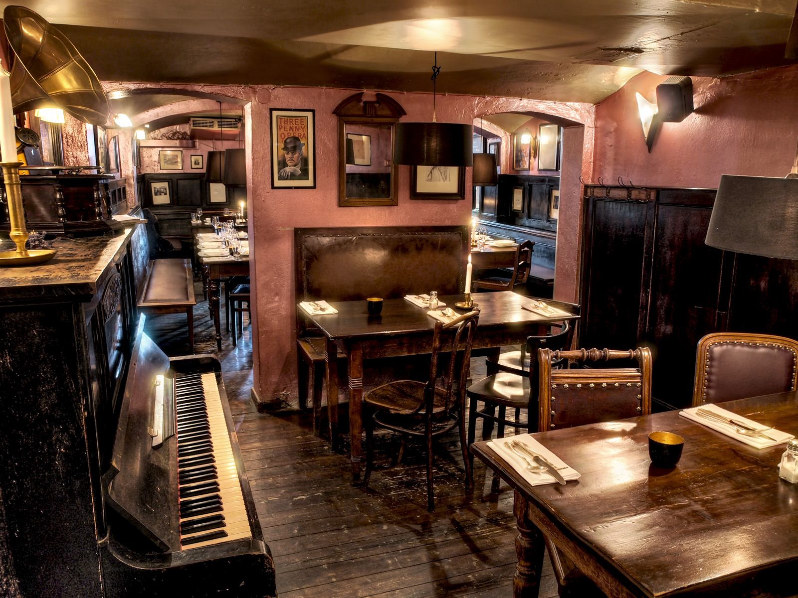 Auf diesem Bild sieht man im Vordergrund ein dunkles Klavier. Dazu einen Tisch aus dunklen Holz, der mit Servietten und Besteck eingedeckt ist. Im Hintergrund befinden sich zahlreiche Bilder an der Wand.