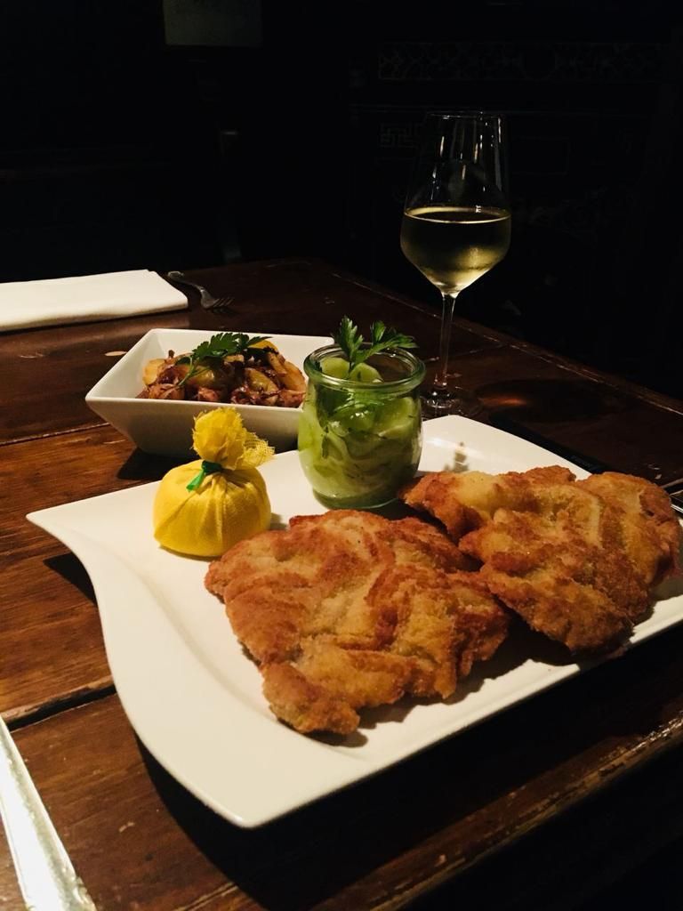 Auf diesem Teller befinden sich zwei appetitlich, braun gebratene Wiener Schnitzel vom Kalb. Dazu ein kleines weg. Glas mit Gurken,salat und eine Zitrone, die in einem kleinen Säckchen verpackt ist. Dahinter befindet sich ein Schälchen mit Bratkartoffeln sowie im anschnitt ein Glas mit Weißwein.