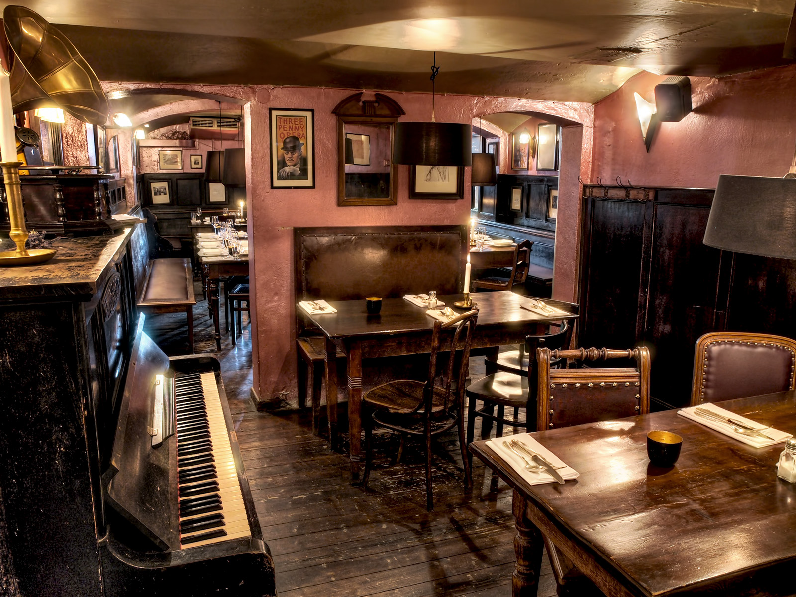 Auf diesem Bild sieht man im Vordergrund ein dunkles Klavier. Dazu einen Tisch aus dunklen Holz, der mit Servietten und Besteck eingedeckt ist. Im Hintergrund befinden sich zahlreiche Bilder an der Wand.